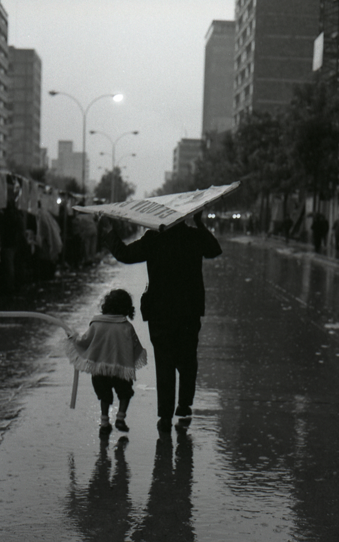 En la fotografía una escena casi que cotidiana, de un adulto y una niña bajo el cielo gris bogotano, resguárdandose de la lluvia. 